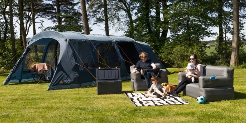Family relaxing in front of a large tent in a grassy area with trees.