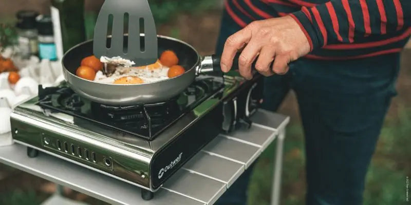 Person cooking outdoors using a portable stove with a frying pan and spatula.