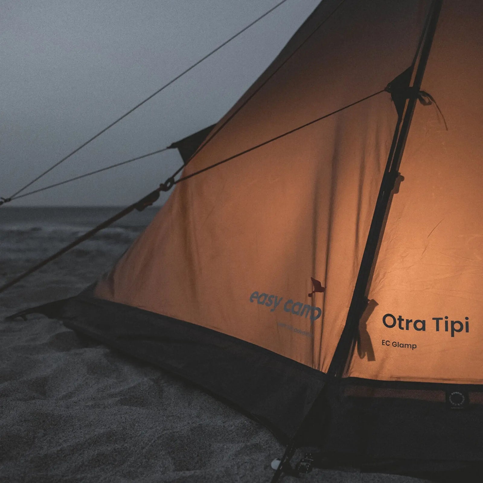 Evening lifestyle shot of the Easy Camp Otra Tipi tent lit from inside, creating a cosy festival atmosphere.