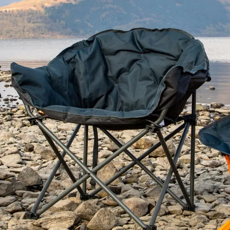 Foldable Vango camping chair with a dark cover on a rocky shore with water and mountains in the background