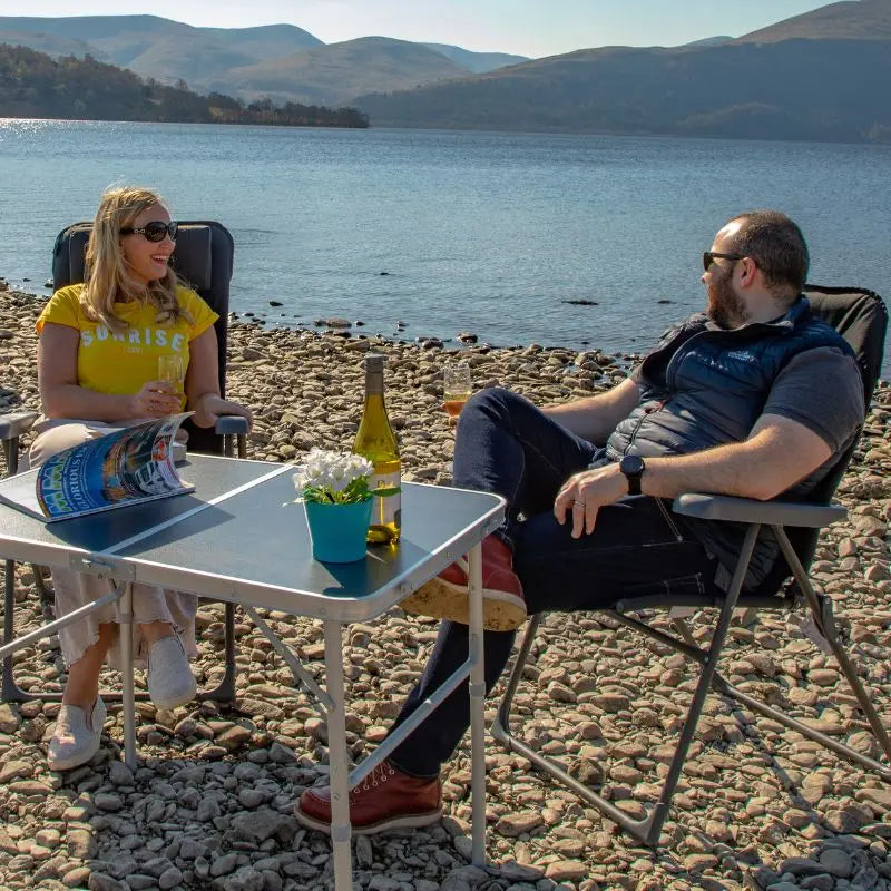 Two people sitting on recling camping chair at a table by a lake, enjoying drinks and a magazine.