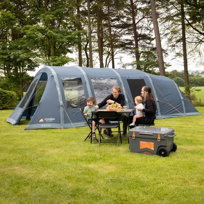 Family camping outdoors with a large Vango tent and cooler in a wooded area.