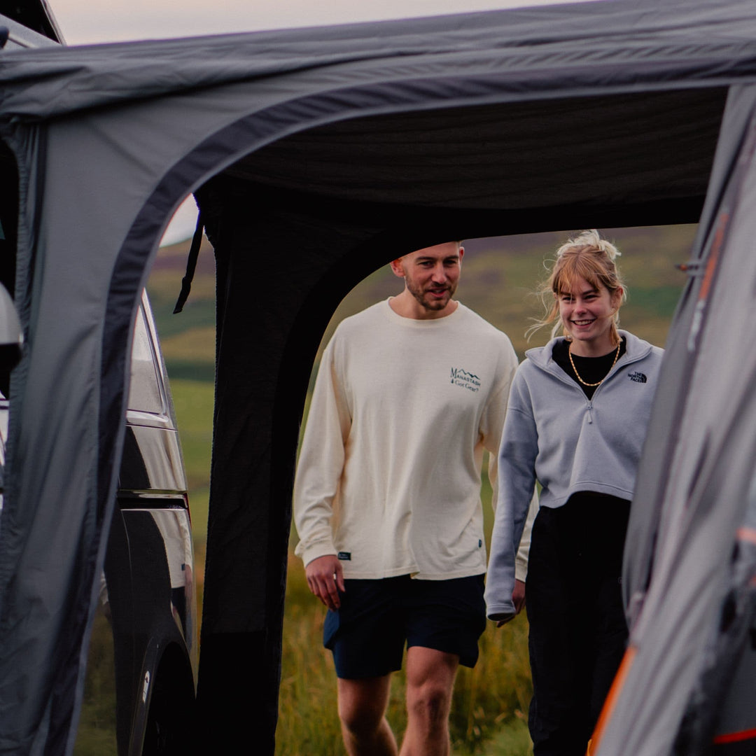 A couple walking through the tunnel area of the Vango Cove III Air Mid Campervan Awning, showing the practical design for outdoor use.