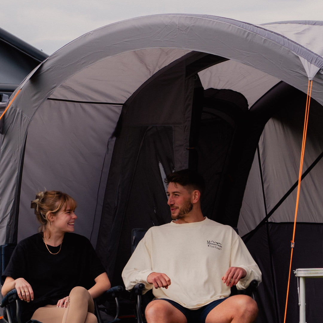 A couple relaxing underneath the sun canopy on the Vango Cove III Inflatable Awning, showing its spacious interior and privacy features with toggled curtains.
