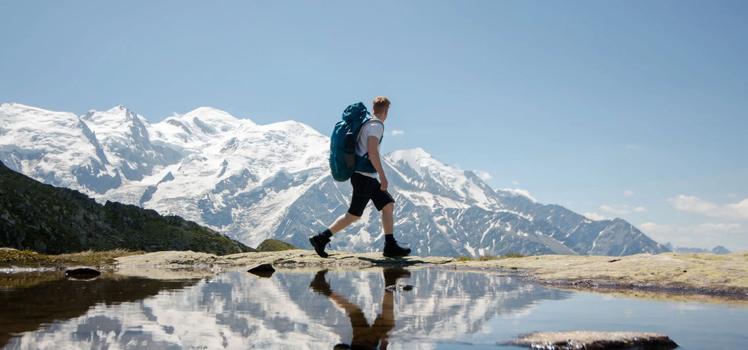 Hiker with a blue backpack walking along a mountain trail with snow-capped peaks reflected in a nearby water pool.