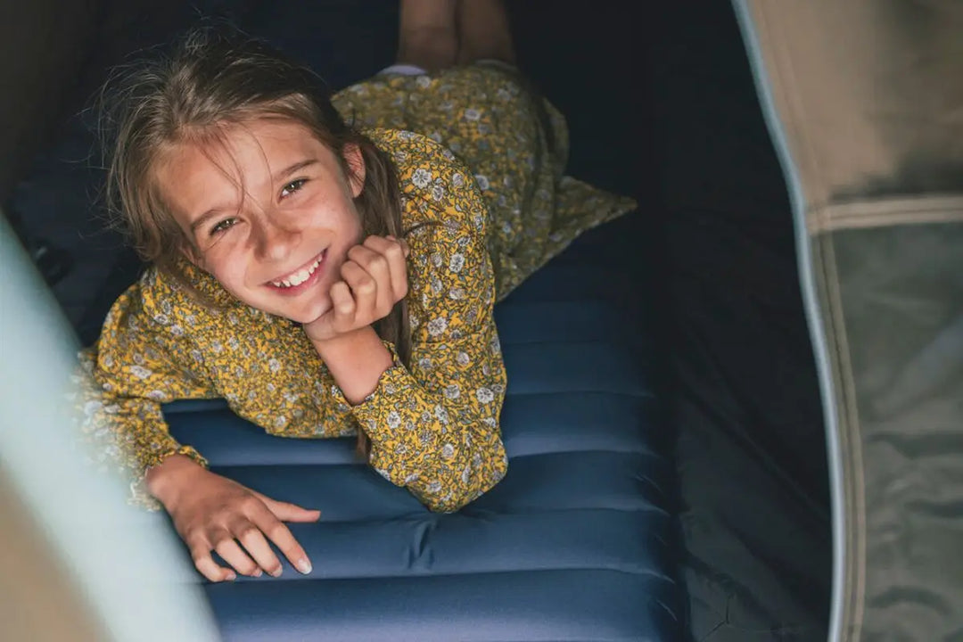 A smiling child lying on a self-inflating mat inside a tent, showcasing comfort and support. The mat is suitable for outdoor camping trips, combining portability with an optimal R-Value for insulation.