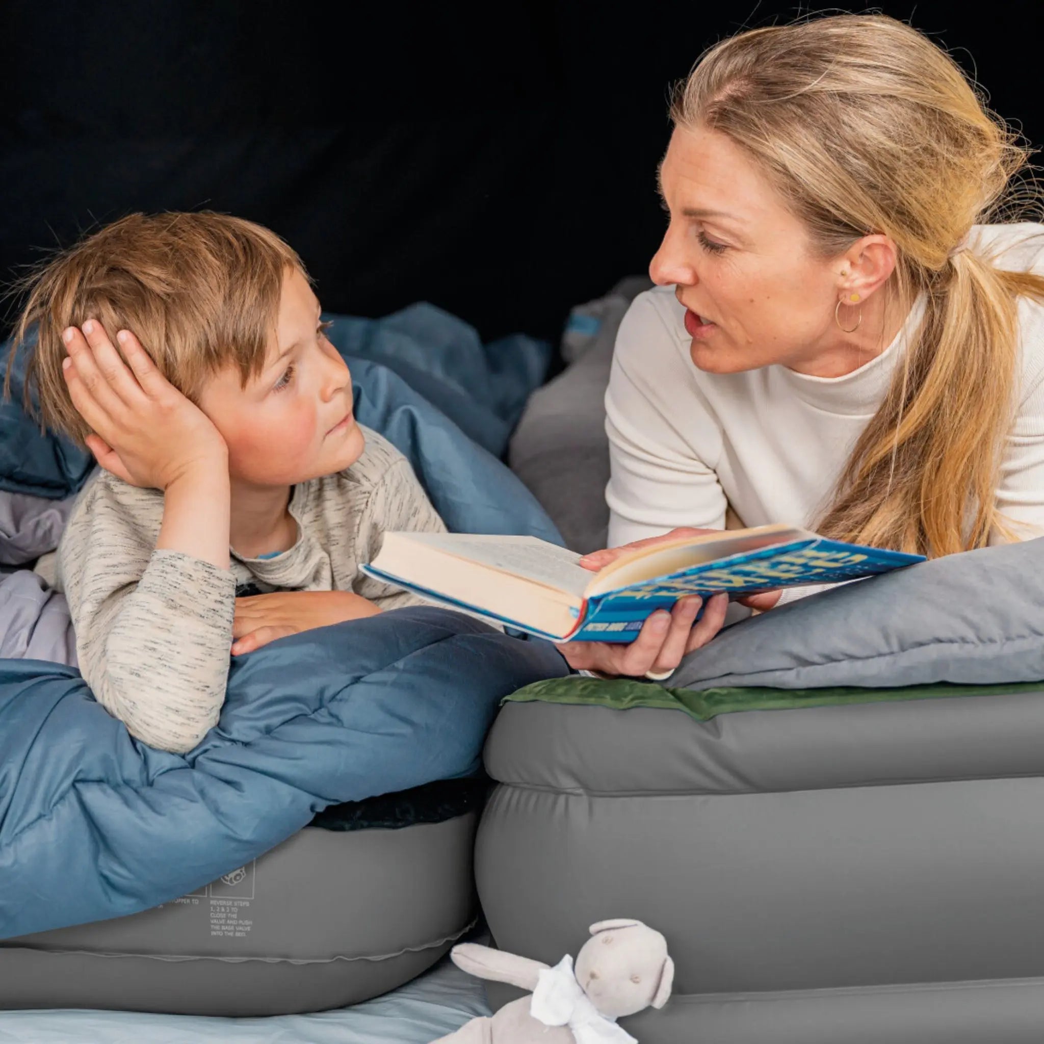 Mother reading to her child while sitting on an inflatable camping bed, ideal for family camping trips.