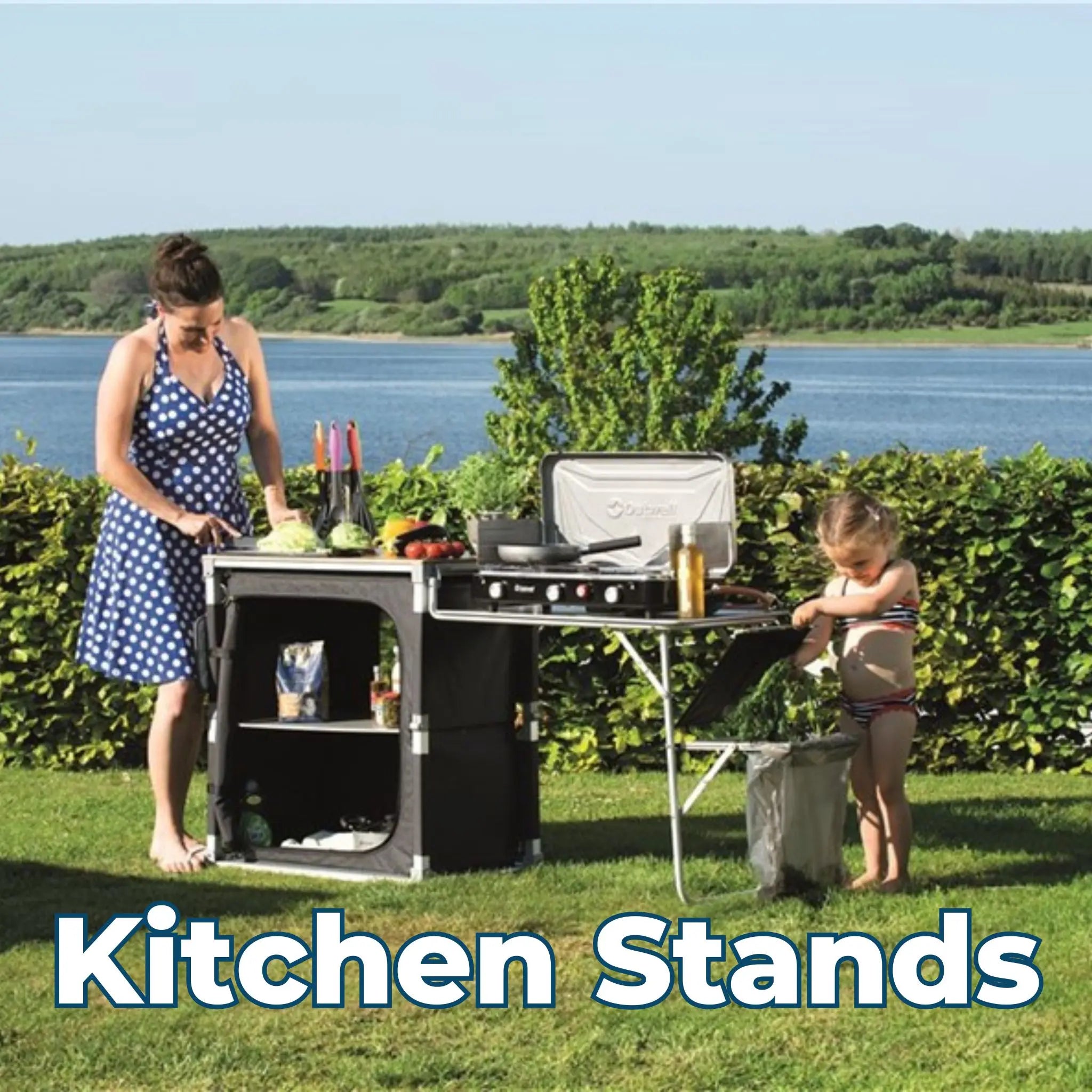 Woman and child using a camping kitchen stand near a lake, preparing food at a portable outdoor cooking station.