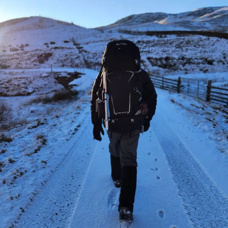 Person with a Vango Duke of Edinburgh rucksack walking on a snow-covered path with mountains in the background.