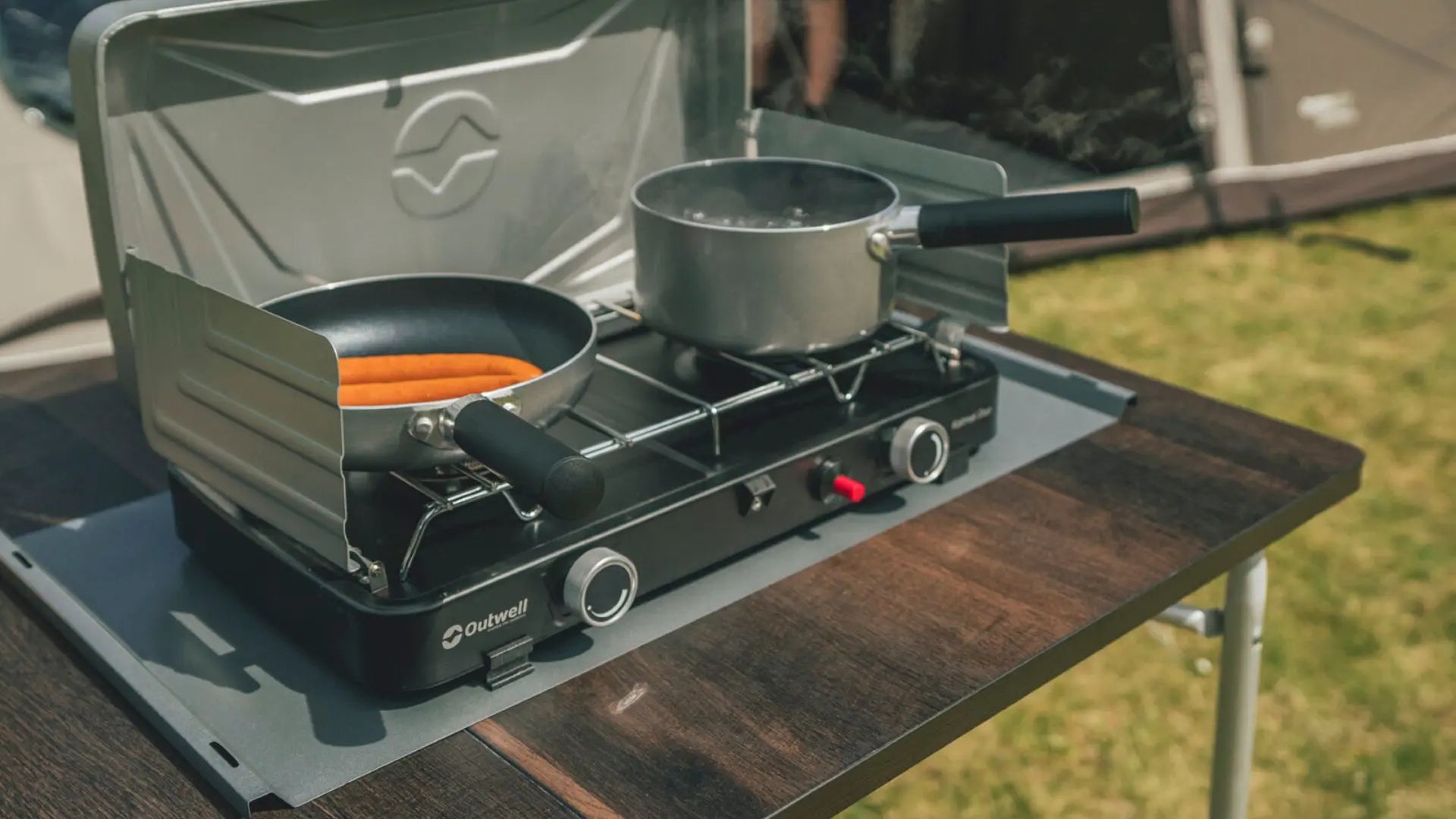 A double burner camping stove on a camping table next to a family tent.