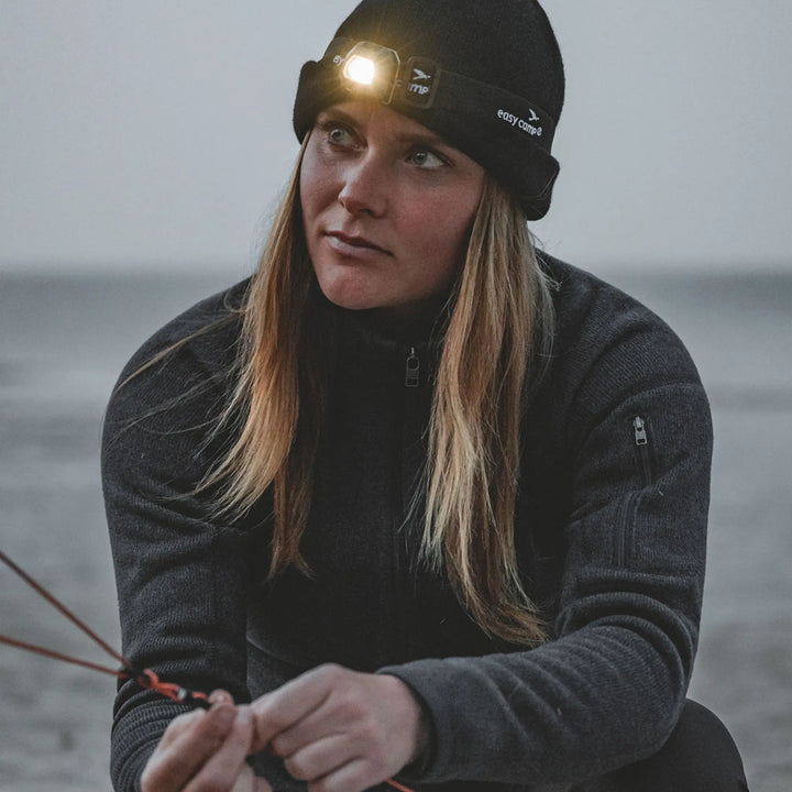 Person wearing a black beanie with a Easy Camp Clover Headlamp, sitting by a body of water.