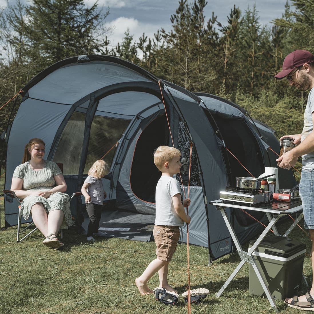 A family enjoying outdoor camping next to the Easy Camp Skarvan 4 tent. A child plays with the guy ropes while a parent prepares food outside.