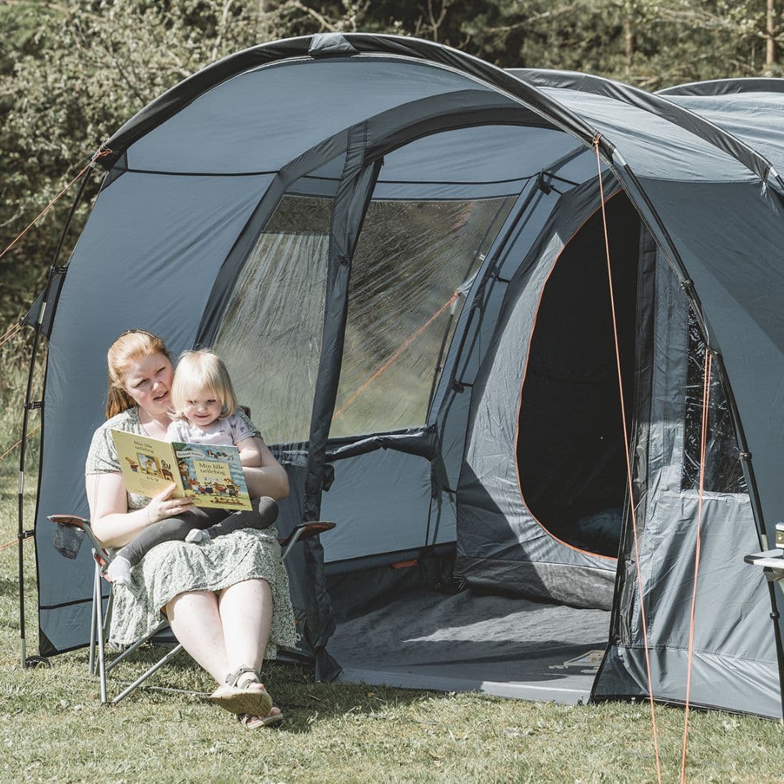 A parent reading a storybook to a child while sitting outside the Easy Camp Skarvan 4 tent in a scenic camping area.