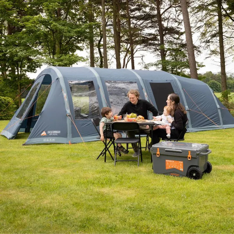 Family camping outdoors with a large inflatable family tent and cooler in a wooded area.