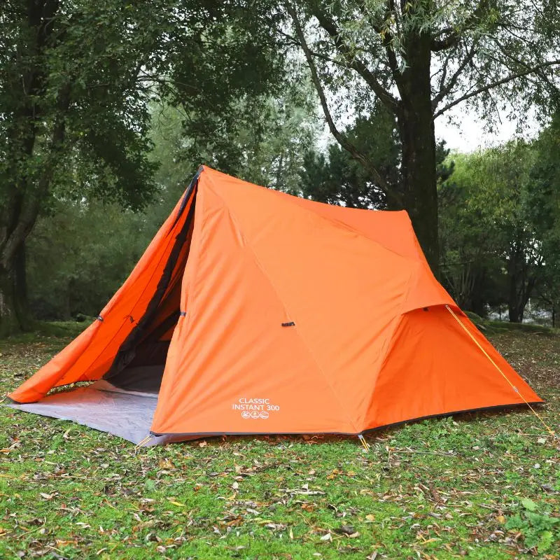 Orange Vango Classic Instant camping tent set up in a grassy area with trees in the background.