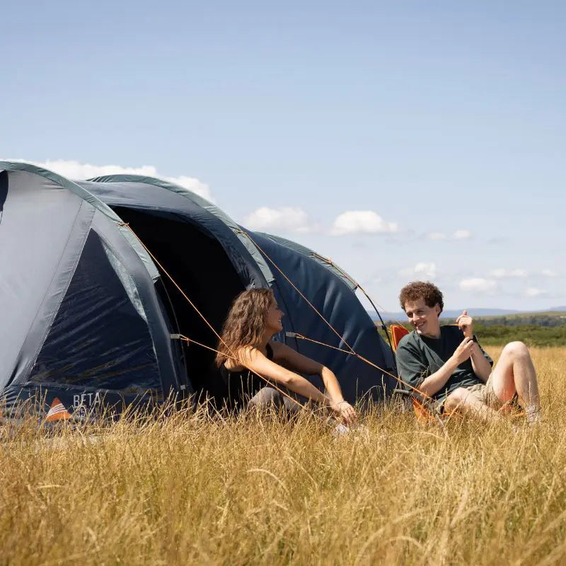 Two people sitting in front of vango Beta 350xl festival tent in a grassy field with a clear sky.