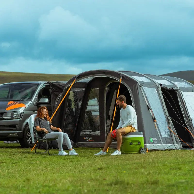 Two people sitting outside a Vango Galli Pro Low inflatable drive away awning in a grassy field.