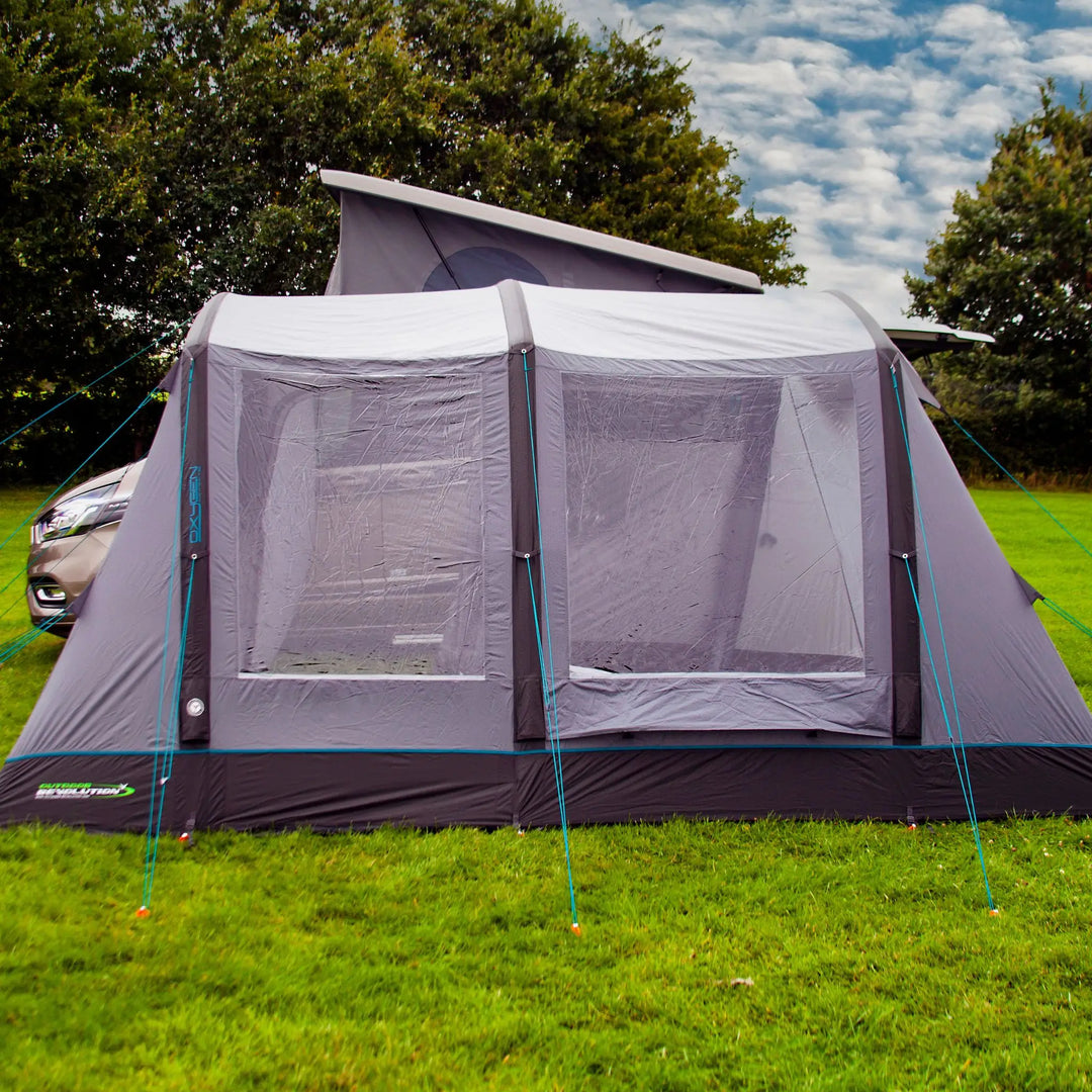 The front of the Outdoor Revolution Cayman Air T3 Low drive away awning, highlighting the spacious entrance, clear windows, and durable inflatable frame, set up beside a campervan.