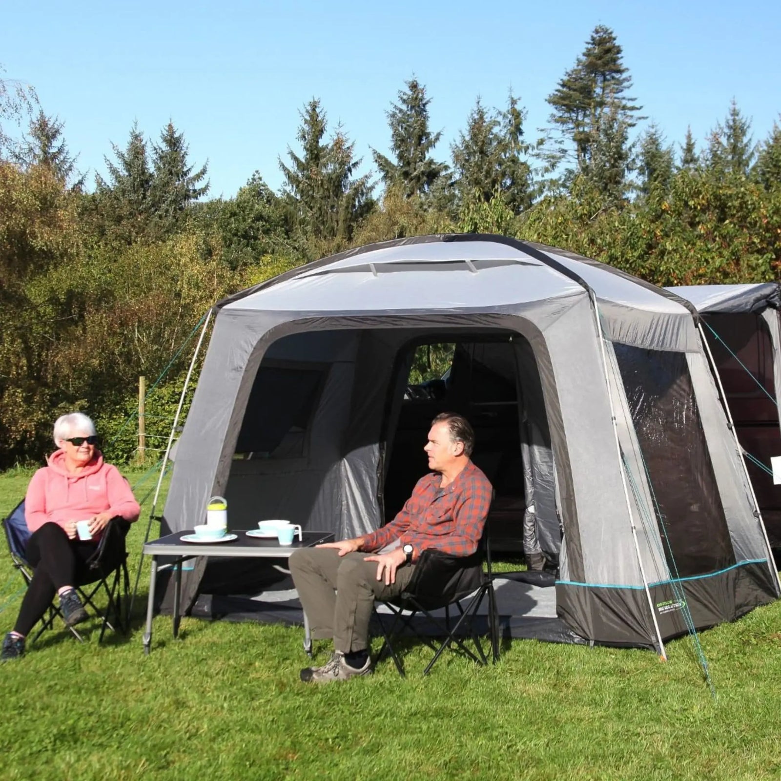 Two people sitting outside a Outdoor Revolution Cayman Cuda DT Awning in a forested area.