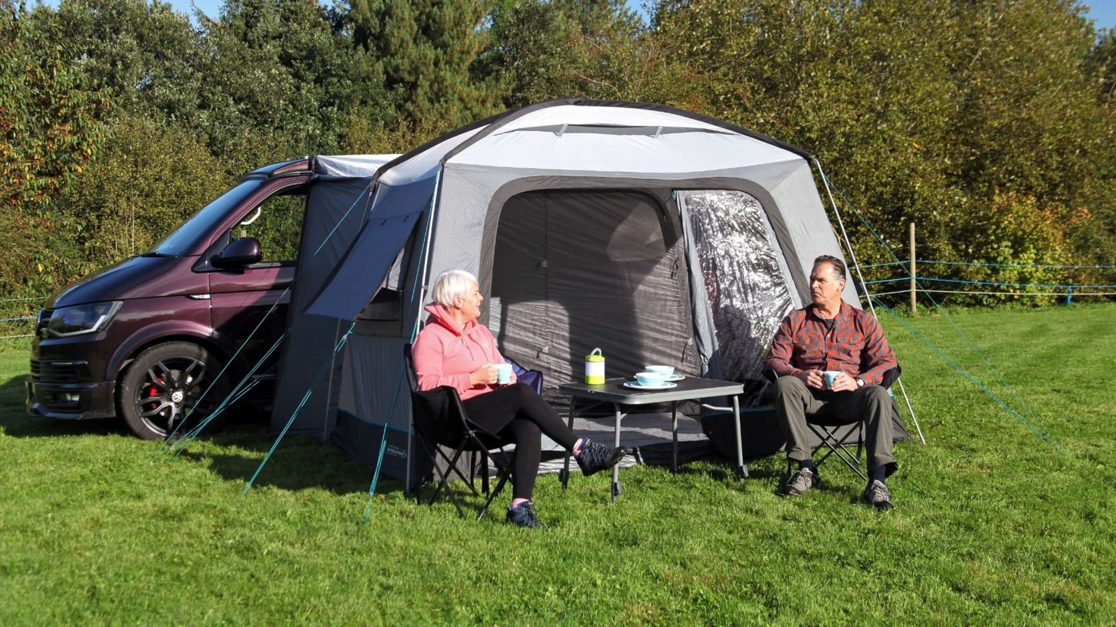 Two people sitting outside the Outdoor Revolution Cayman Cuda F/G Drive Away Awning, enjoying a sunny day next to their campervan. The fibreglass drive away awning is attached to the campervan, showcasing its versatility and spacious design. The Cayman Cuda fibreglass awning provides a functional outdoor living space, perfect for camping trips.