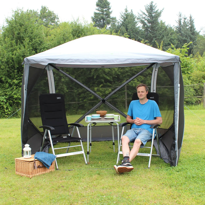 Man sitting inside an Outdoor Revolution Screenhouse 4 shelter on a camping chair, with a table and picnic accessories.