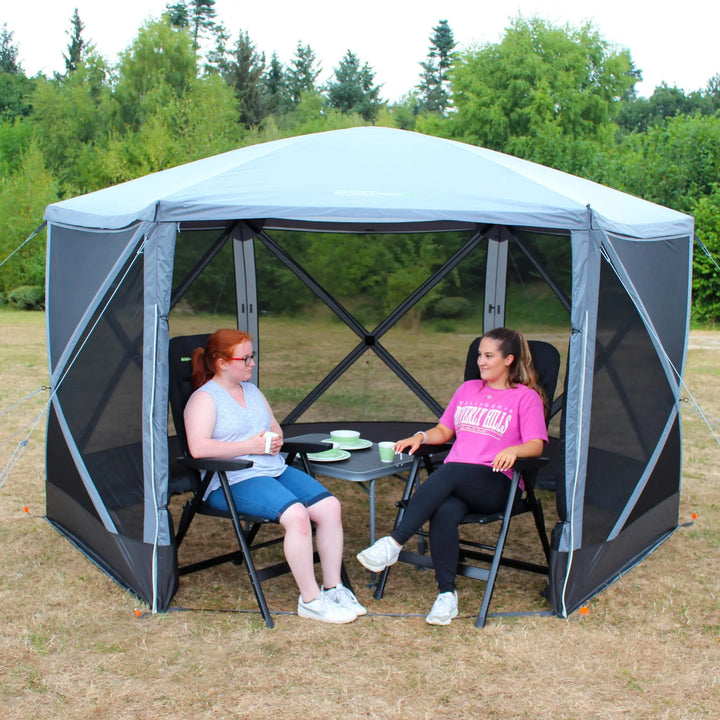 Two women sitting inside an Outdoor Revolution Screenhouse 6 shelter, enjoying drinks at a table in a grassy outdoor setting.