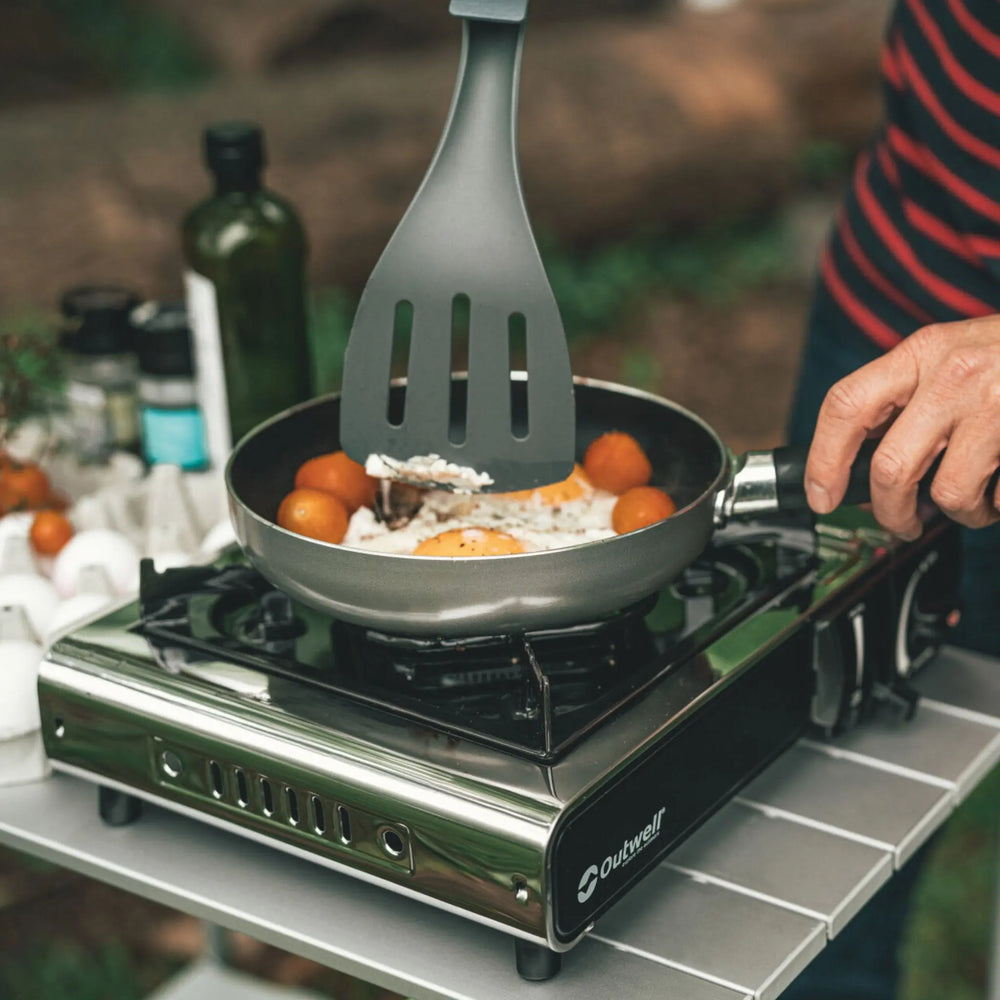 A person cooking eggs and tomatoes in a pan on the Outwell Appetizer Solo Stove, placed on a camping table outdoors.