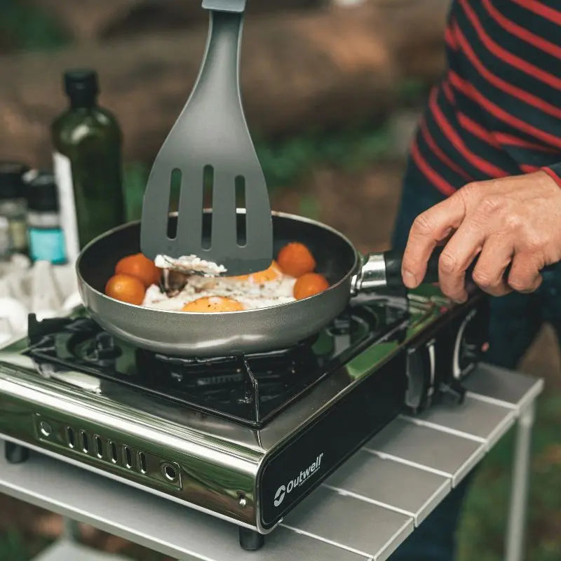 Person cooking outdoors using an Outwell stove with a frying pan and spatula.