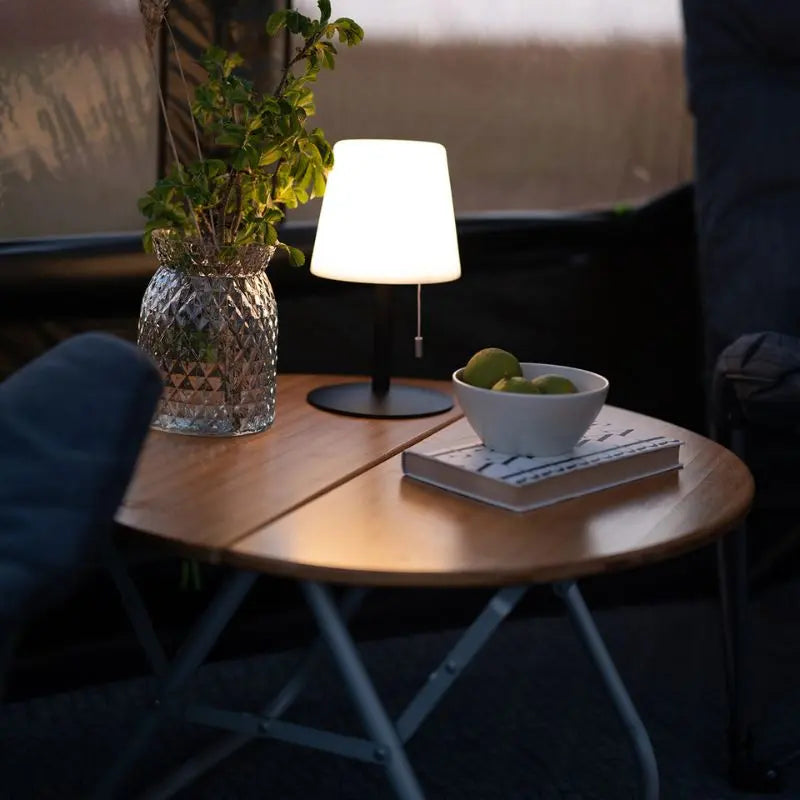 Small wooden table with an Outwell lamp, vase, and bowl of fruit in a dimly lit room.