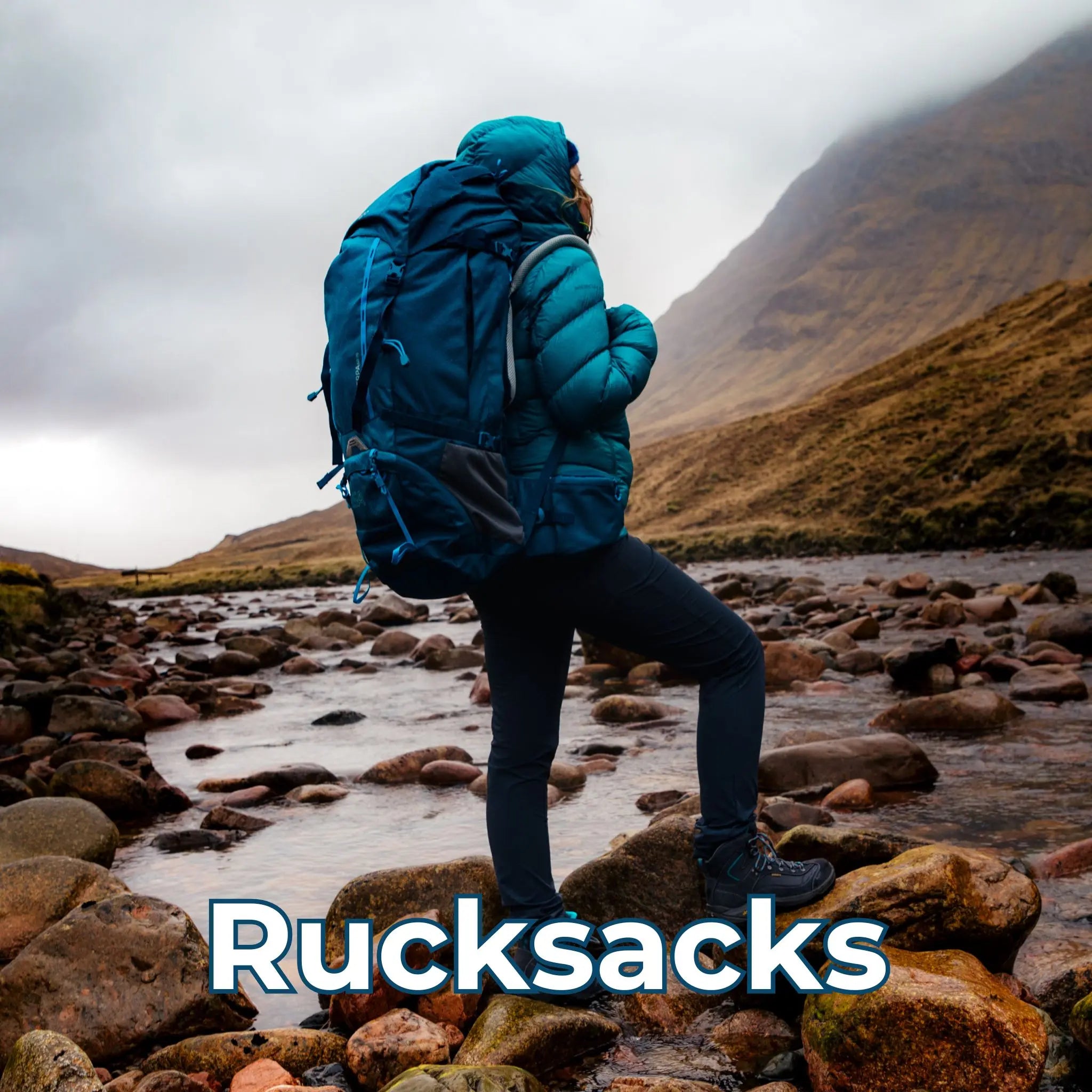 Rucksacks – A hiker wearing a large blue rucksack and a matching jacket, trekking through a rocky riverbed with mountains in the background.