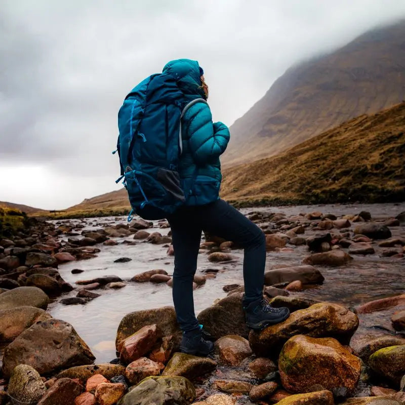 Person in a blue jacket and rucksack standing on rocks by a stream with mountains in the background.