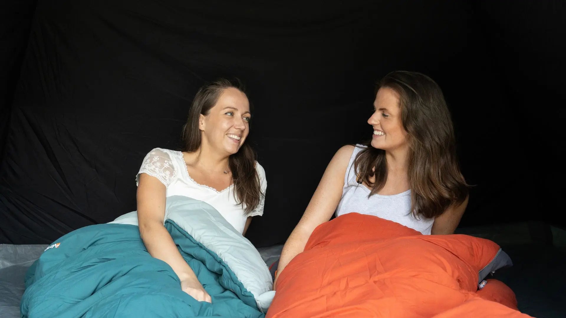 Two women sitting on a bed with teal and orange sleeping bags in a tent.