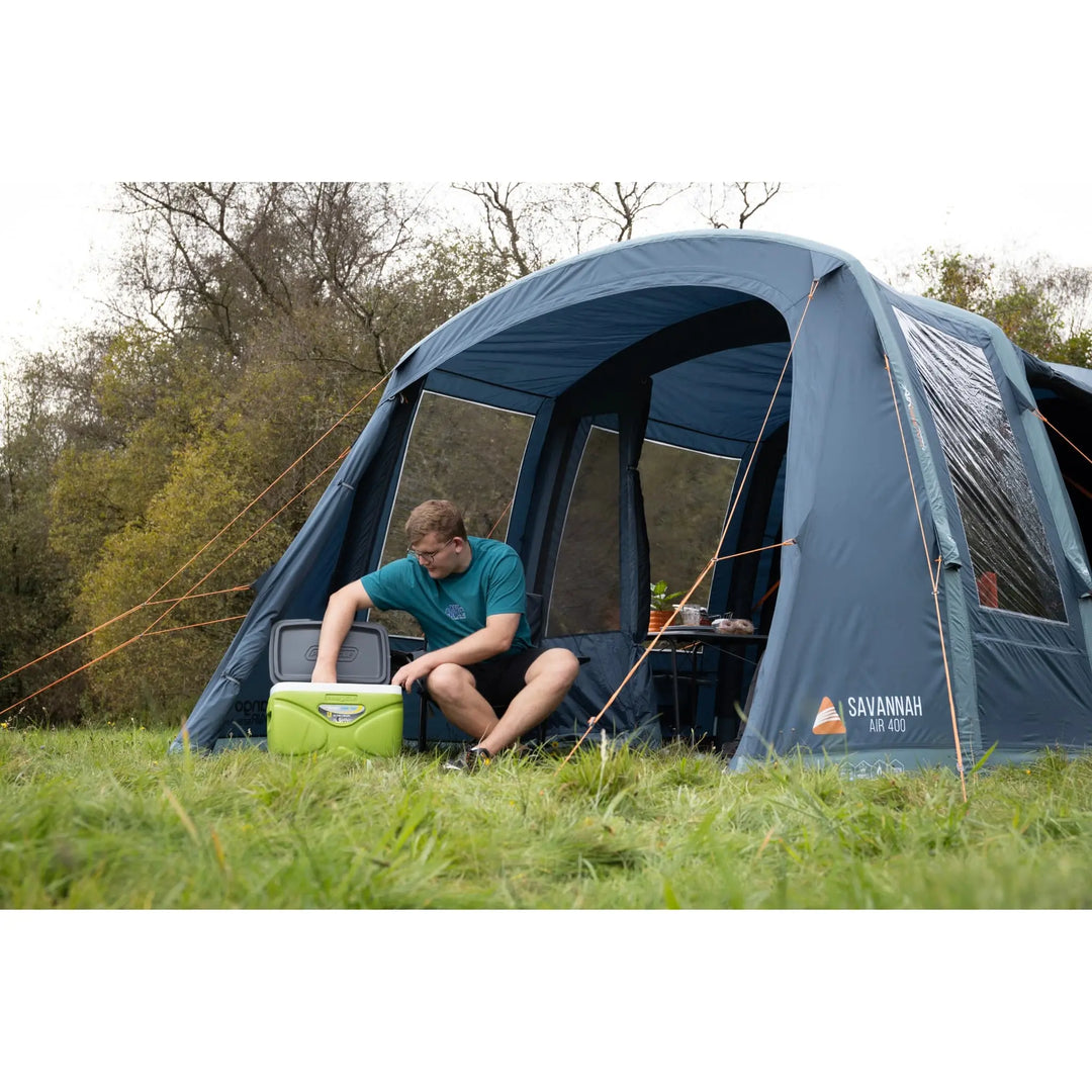 Camper sitting outside the Vango Savannah Air 400 large camping tent in a grassy field, using a cooler—perfect for glamping, festivals, and outdoor adventures.