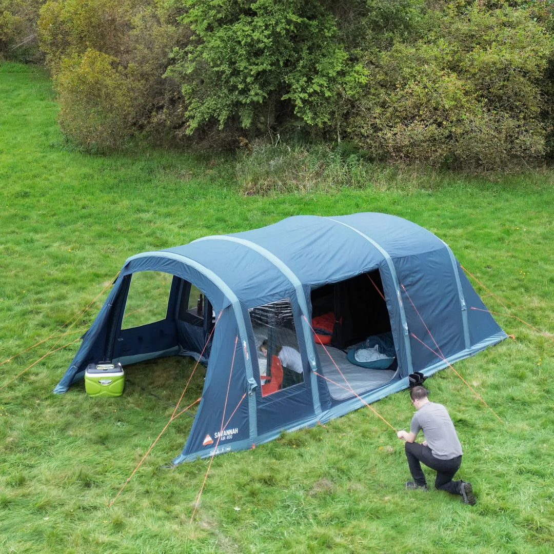 Aerial view of the Vango Savannah Air 400 family tent pitched on a grassy field, with a camper securing guy lines—ideal for weekend camping trips and outdoor adventures.