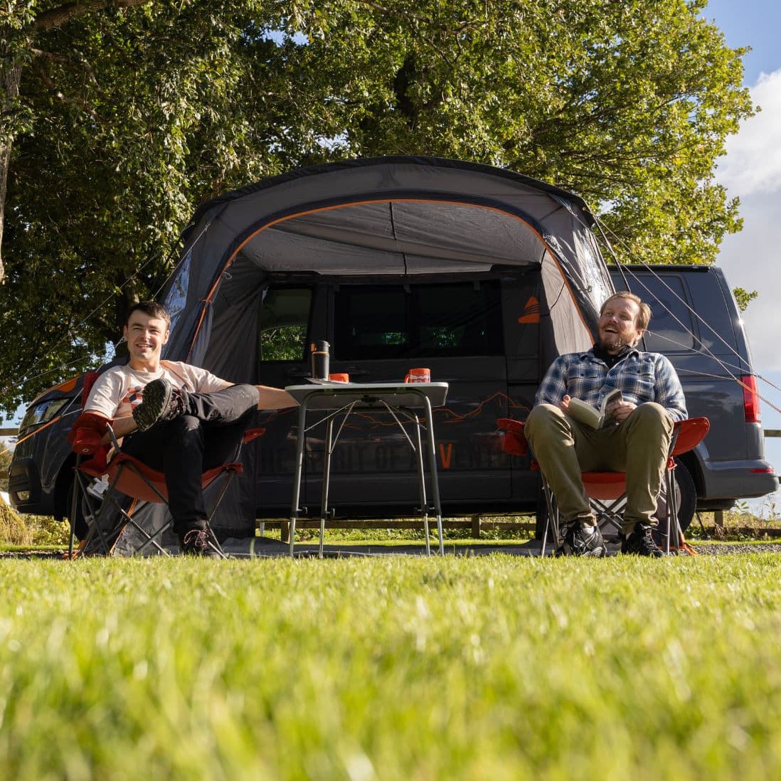 Lifestyle image of two people relaxing outside the Vango Arden Tailgate Awning, with the VW campervan parked at a scenic campsite.