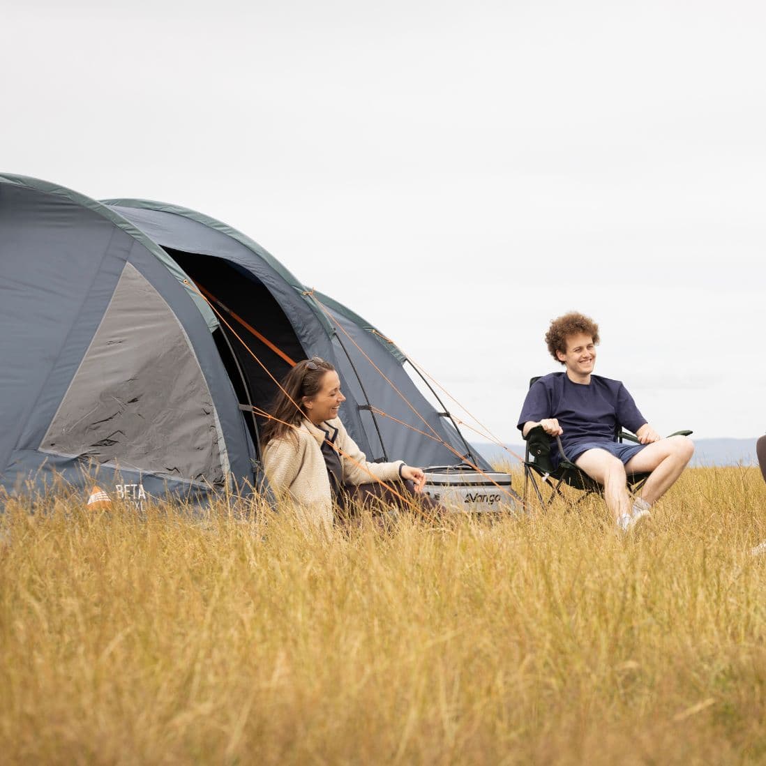 Couple sitting outside the Vango Beta 450XL tent in a grassy field, enjoying a relaxed camping setup.