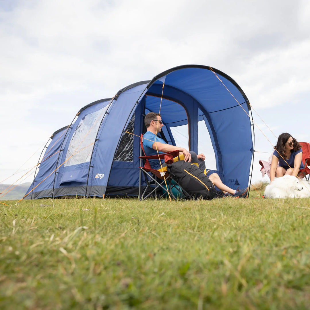 Two people sitting outside a Vango Capri 400 Tent in a grassy field with a cloudy sky.