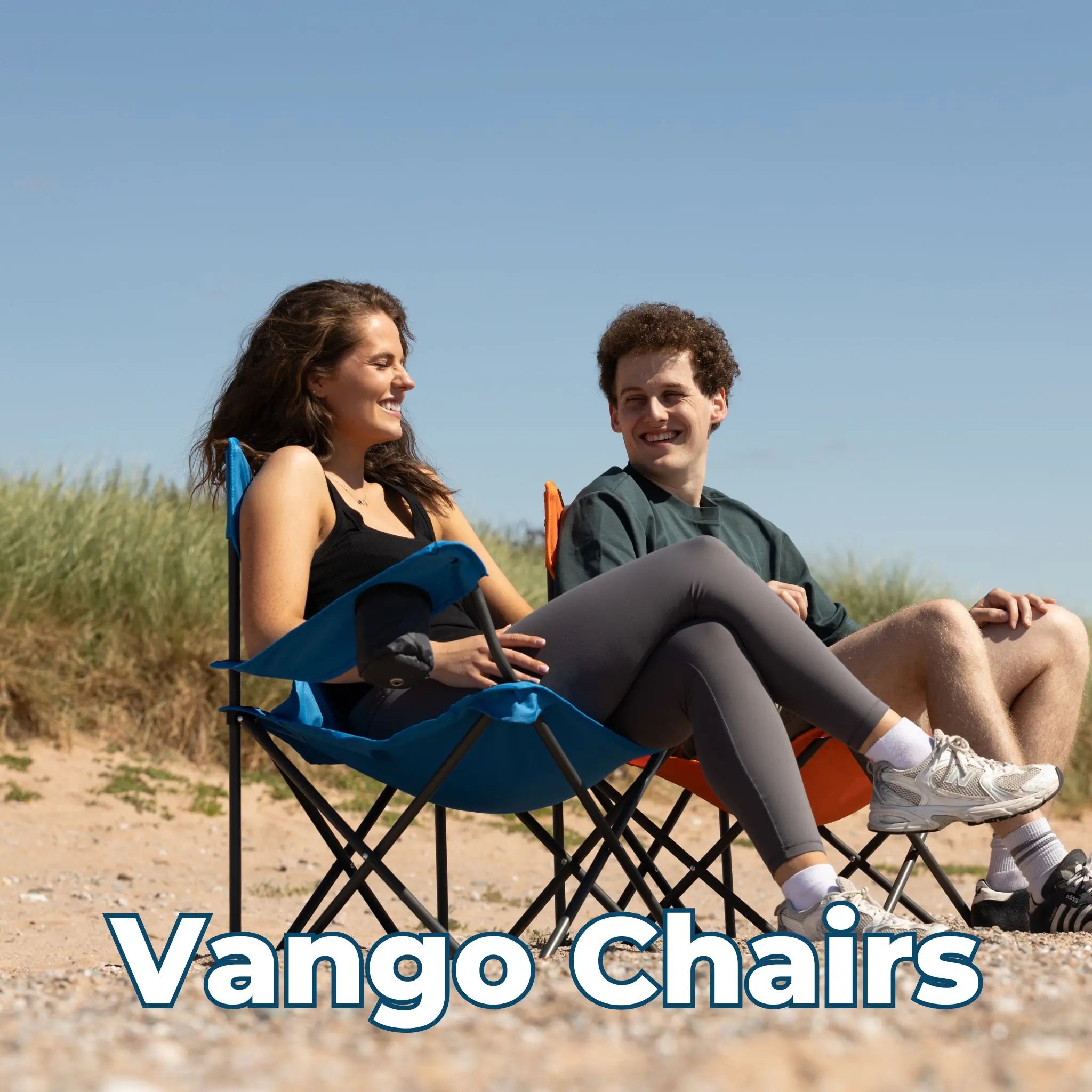 Two people relaxing on Vango camping chairs on a beach, sitting in foldable blue and orange chairs under a clear sky.