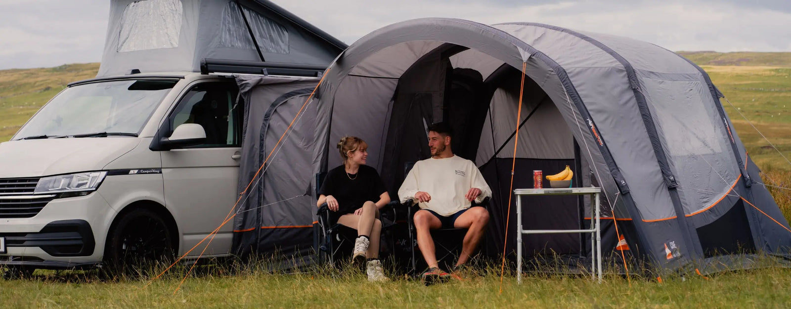 A campervan with a Vango Cove III Air Low Drive Away Awning attached, featuring two people seated comfortably at the entrance with a table holding refreshments nearby, set in a scenic countryside.