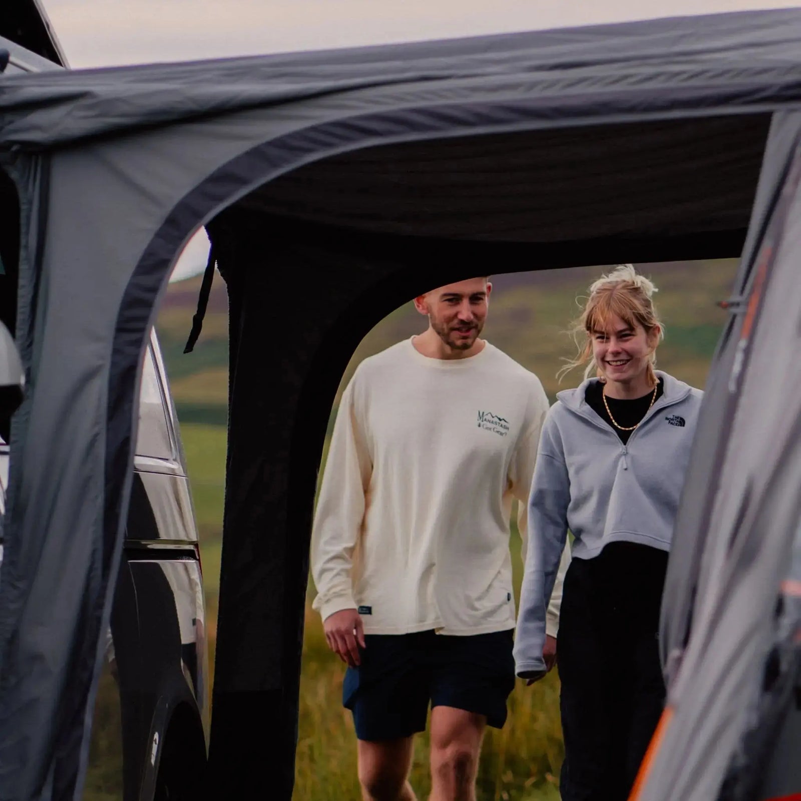 Two people walking under the sheltered connection tunnel between a campervan and the Vango Cove III Air Low Drive Away Awning, providing a glimpse of the convenient drive-away setup.