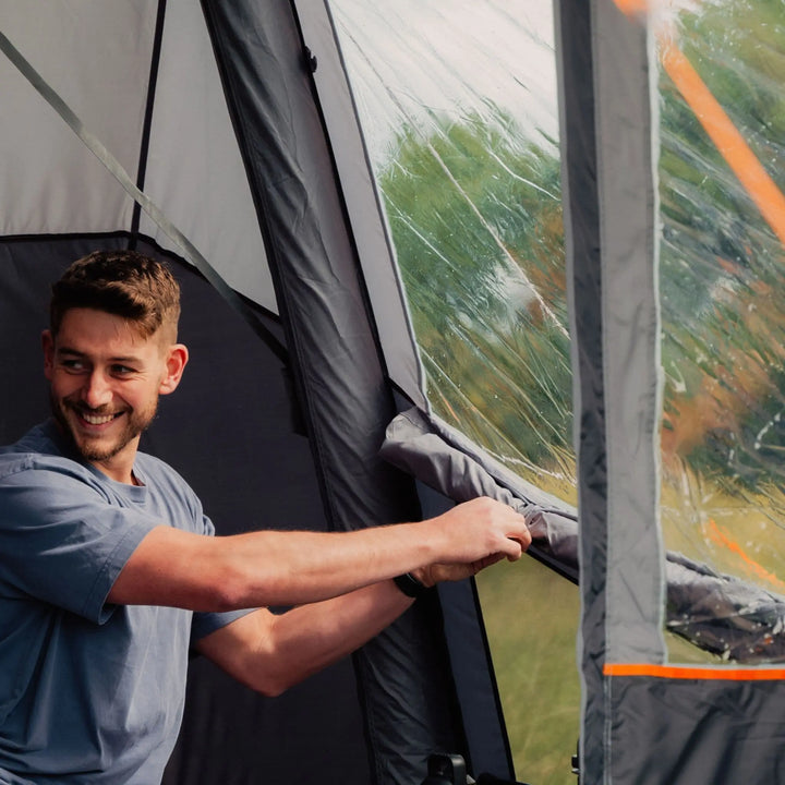 Man rolling up toggled privacy curtains inside the Vango Cove III Air Low Drive Away Awning, showcasing interior space and Diamond Clear windows.