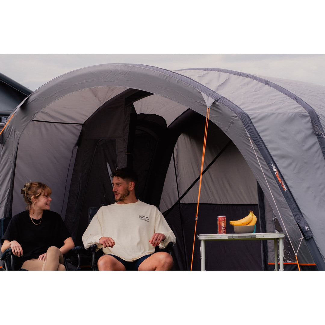 A couple relaxing under the canopy of the Vango Cove III Air Mid Inflatable Awning, highlighting its versatile outdoor living area.