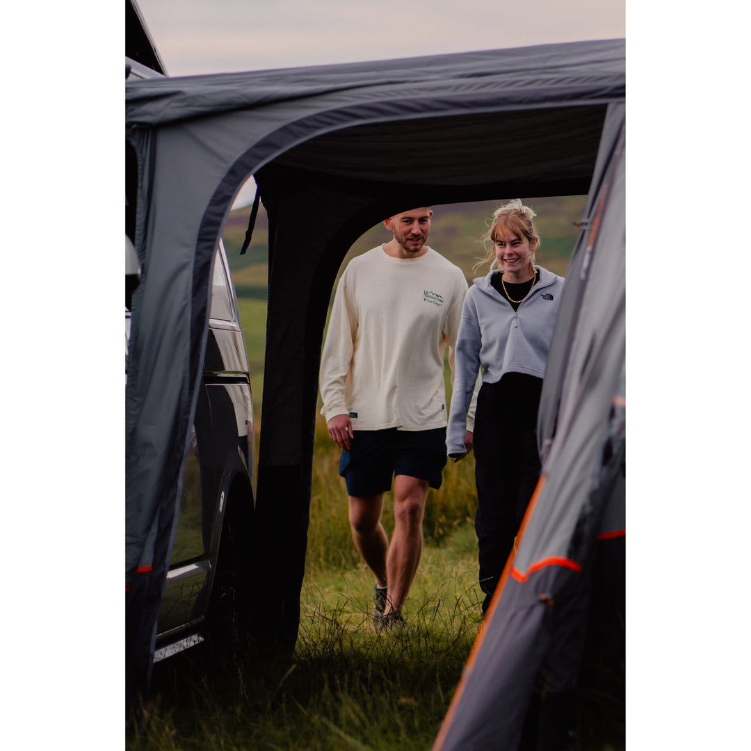 A couple walking through the connecting tunnel of the Vango Cove III Air Mid Drive Away Awning, illustrating its easy attachment to campervans and motorhomes for extra living space on camping trips.