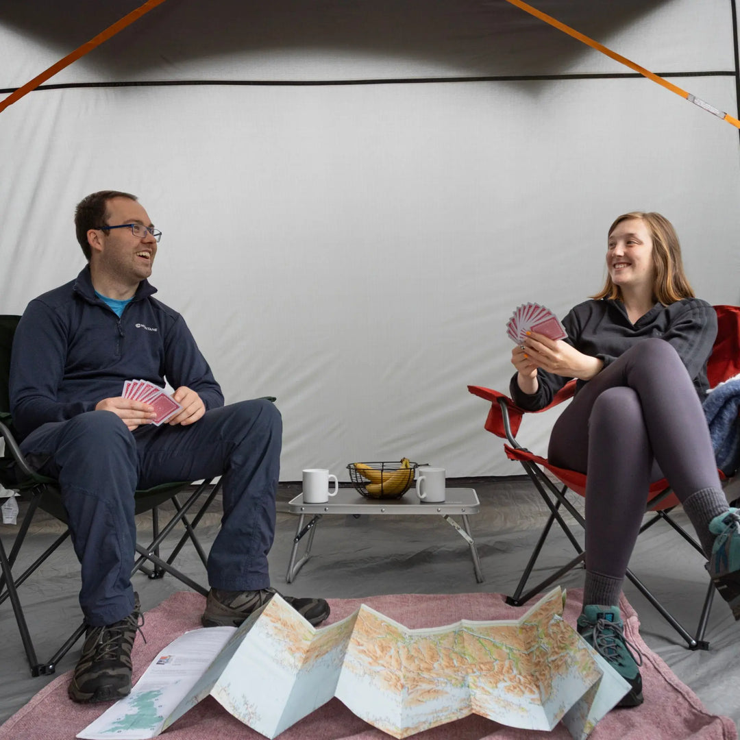 A man and a woman sitting in folding camp chairs inside a Vango Cove III Mid (Poled) Awning 2026, smiling and playing with maps and cards on a small table between them.