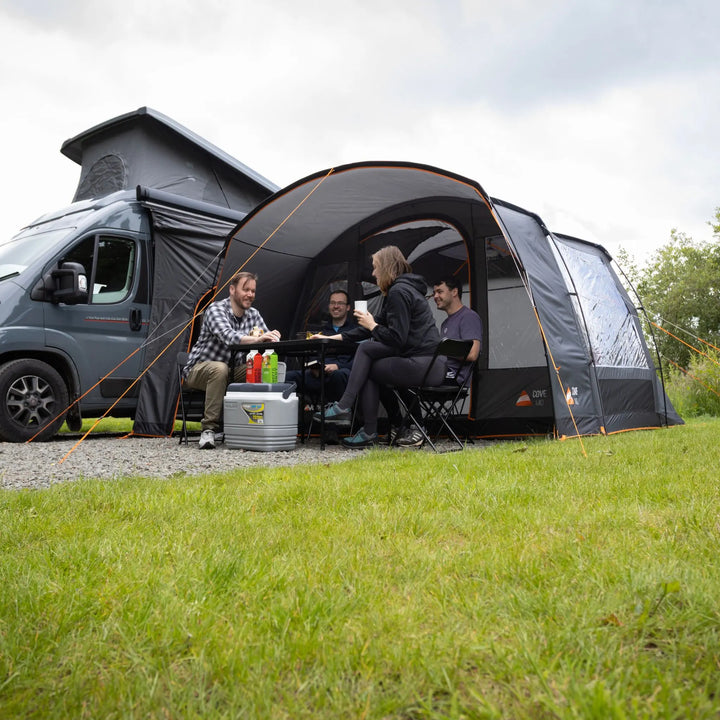 A Vango Cove III Mid (Poled) Awning 2026 is pitched beside a gray motorhome on a grassy campsite. Three people are seated inside the tent area with a small table and cooler, and trees are visible in the background under an overcast sky.