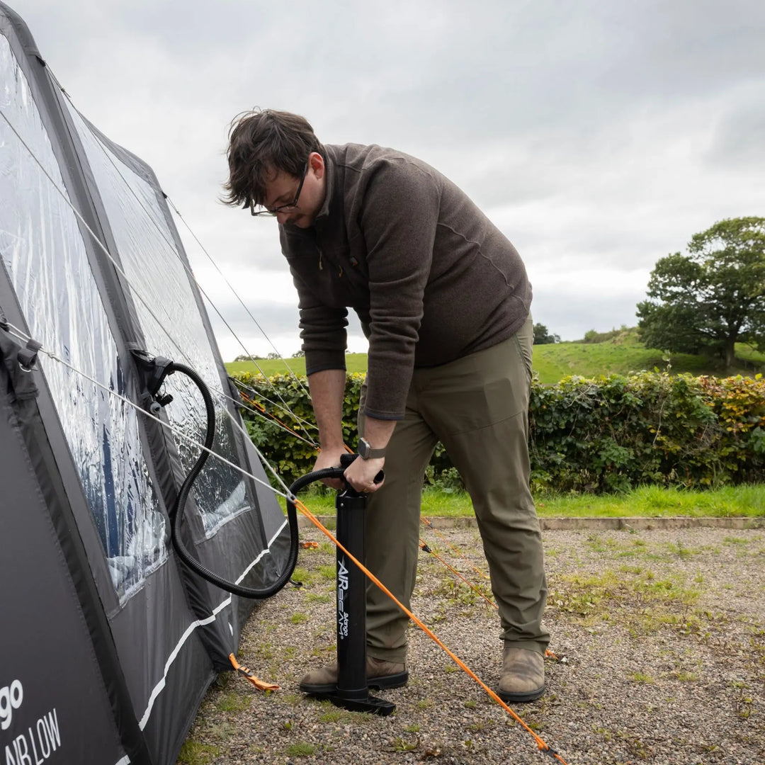 A man outdoors using a manual air pump to inflate a Vango Cove Pro Air Low Drive Away Awning, set against a grassy field under an overcast sky.