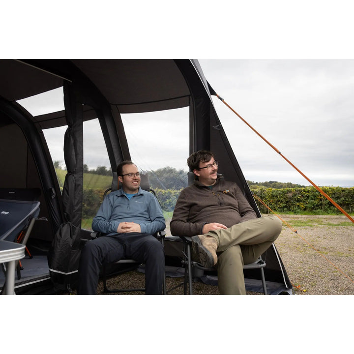Two men sitting in camping chairs inside a Vango Cove Pro Air Low Drive Away Awning, with an outdoor grassy landscape visible through the open sides under an overcast sky.