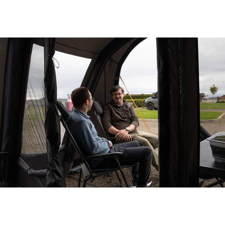 Two men sitting in folding chairs inside the front porch of the Vango Cove Pro Air Low Drive Away Awning, with an outdoor lawn and cloudy sky visible through the open front door.