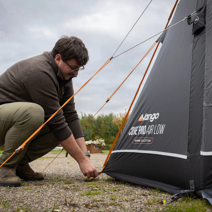 A man crouches outdoors to attach a tent stake to a Vango Cove Pro Air Low Drive Away Awning on a gravel surface under an overcast sky.