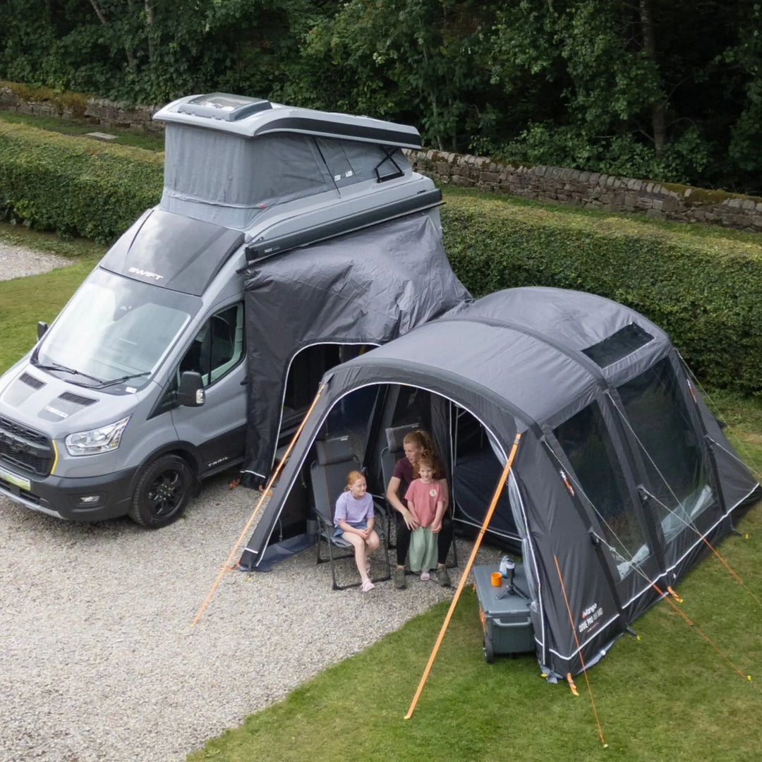 A grey pop-top camper van parked outdoors on a gravel driveway beside a Vango Cove Pro Air Mid Drive-Away Awning with two people sitting inside, with green hedges and trees in the background.